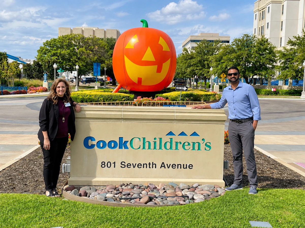 Two people standing on either side of the Cook Children’s sign, with a large pumpkin display behind them
